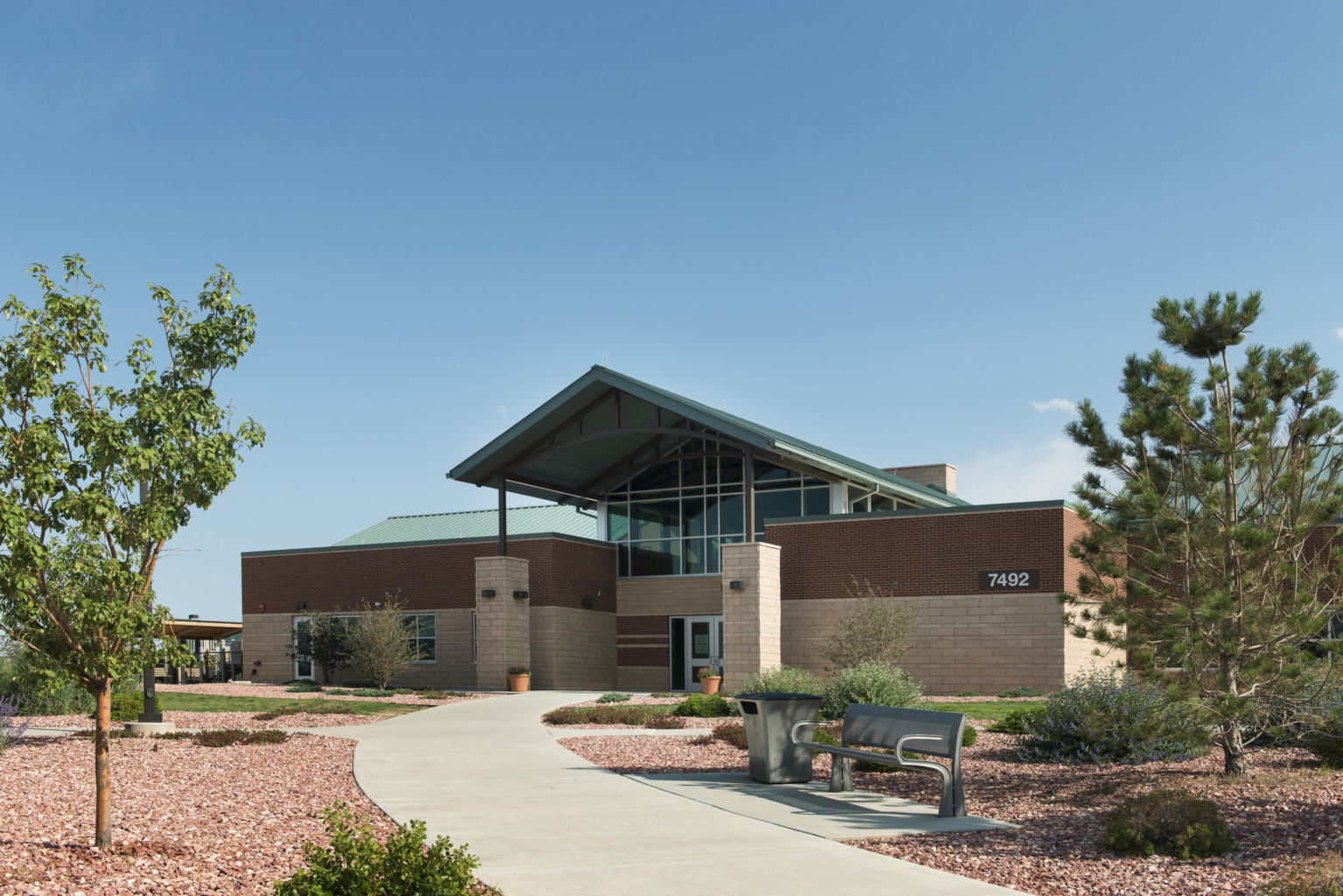 A view of Warrior in Transition complex with two red and pink brick buildings joined by glass atrium, curved stone path through landscaping to peaked blue entry canopy