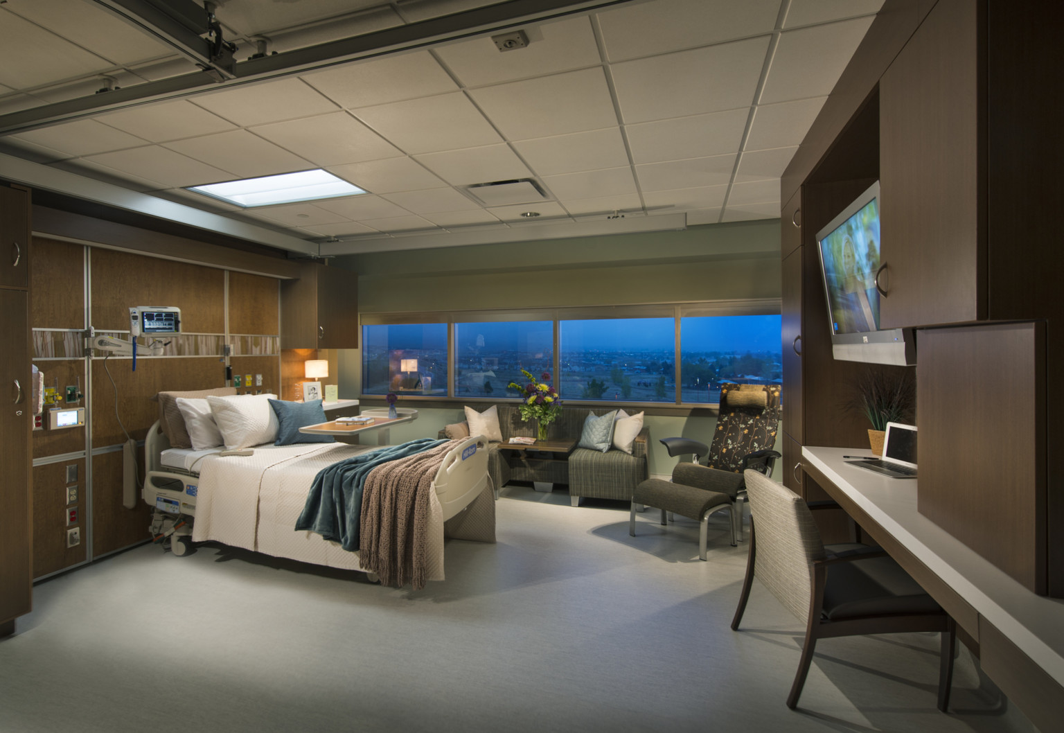 patient room with wood finish walls, floral and neutral colored upholstered guest seating. Exterior wall lined with windows
