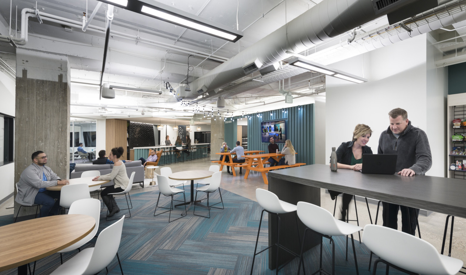 employee lounge area with tvs, picnic benches, standing tables in white room with exposed ducts