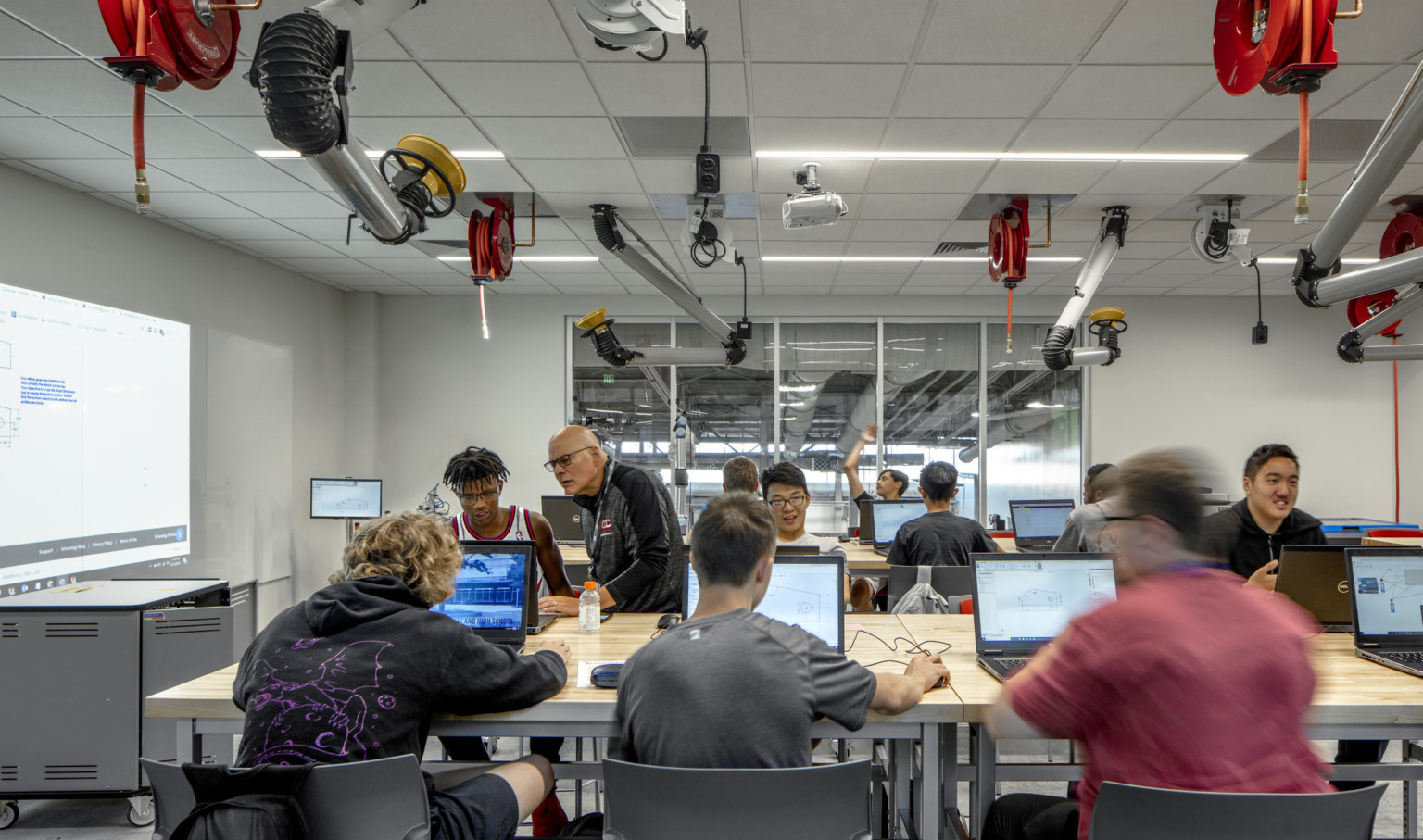 Classroom with students on computers at rows of wood tables. Equipment and power cords hang from the ceiling of white room