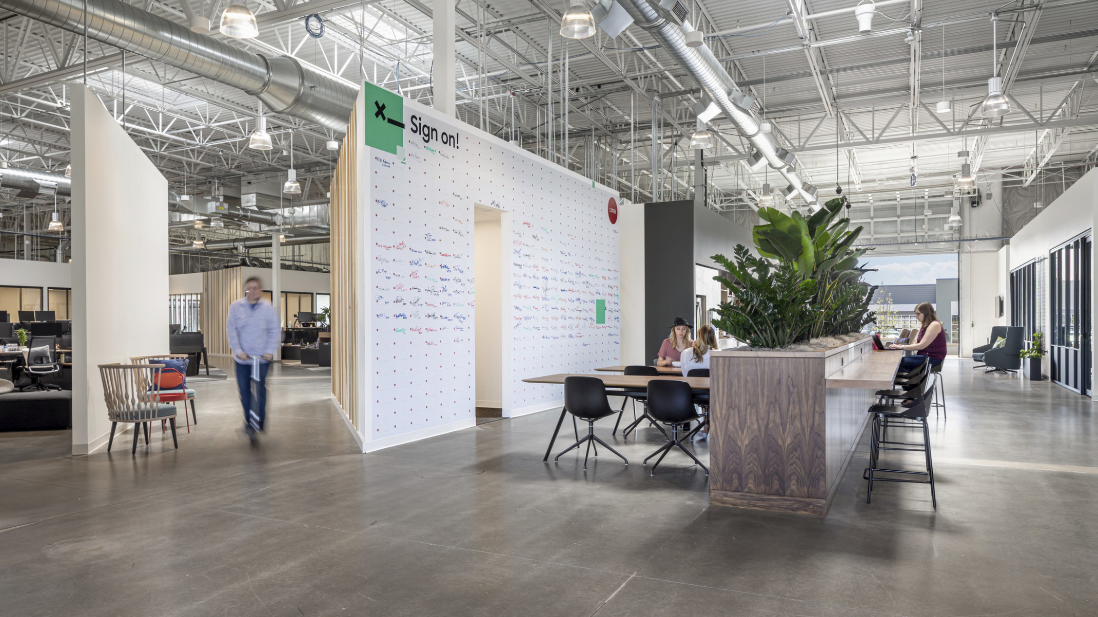 Tables in front of wood half wall with plants on top and counter attached. White board wall to left. Open garage door window
