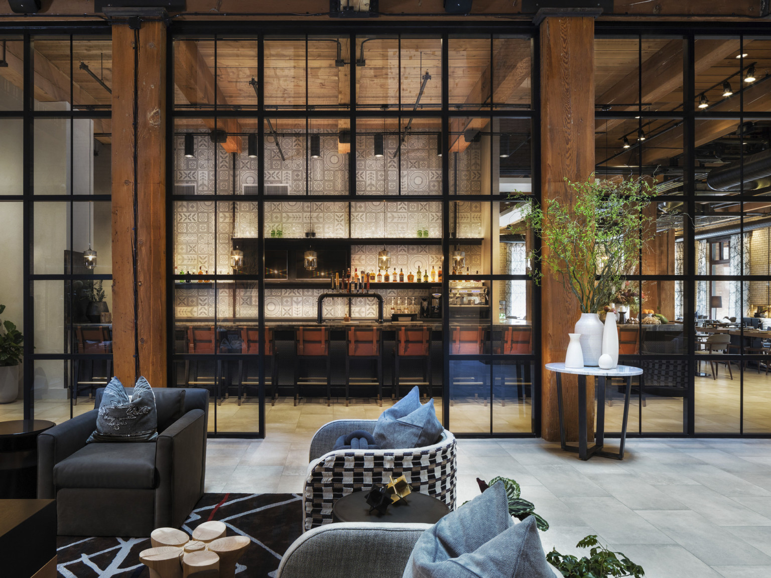 Seating area in room with double height windows looking to white geometric walled bar in next room. Wood columns and ceiling