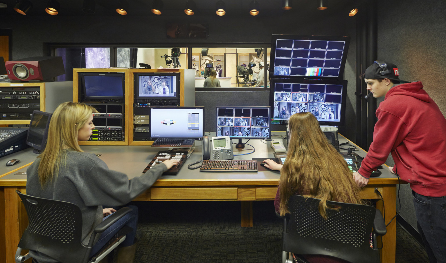 students working in recording studio with multiple screens and audio equipment. Small window above looks into recording booth