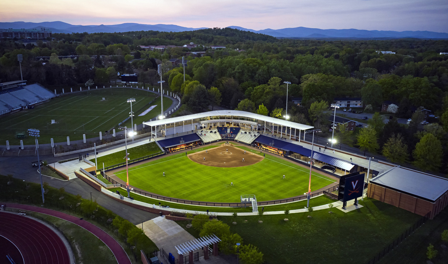 University of Virginia Softball stadium aerial view at lit up at dusk, surrounded by trees and view of Blue Ridge Mountains