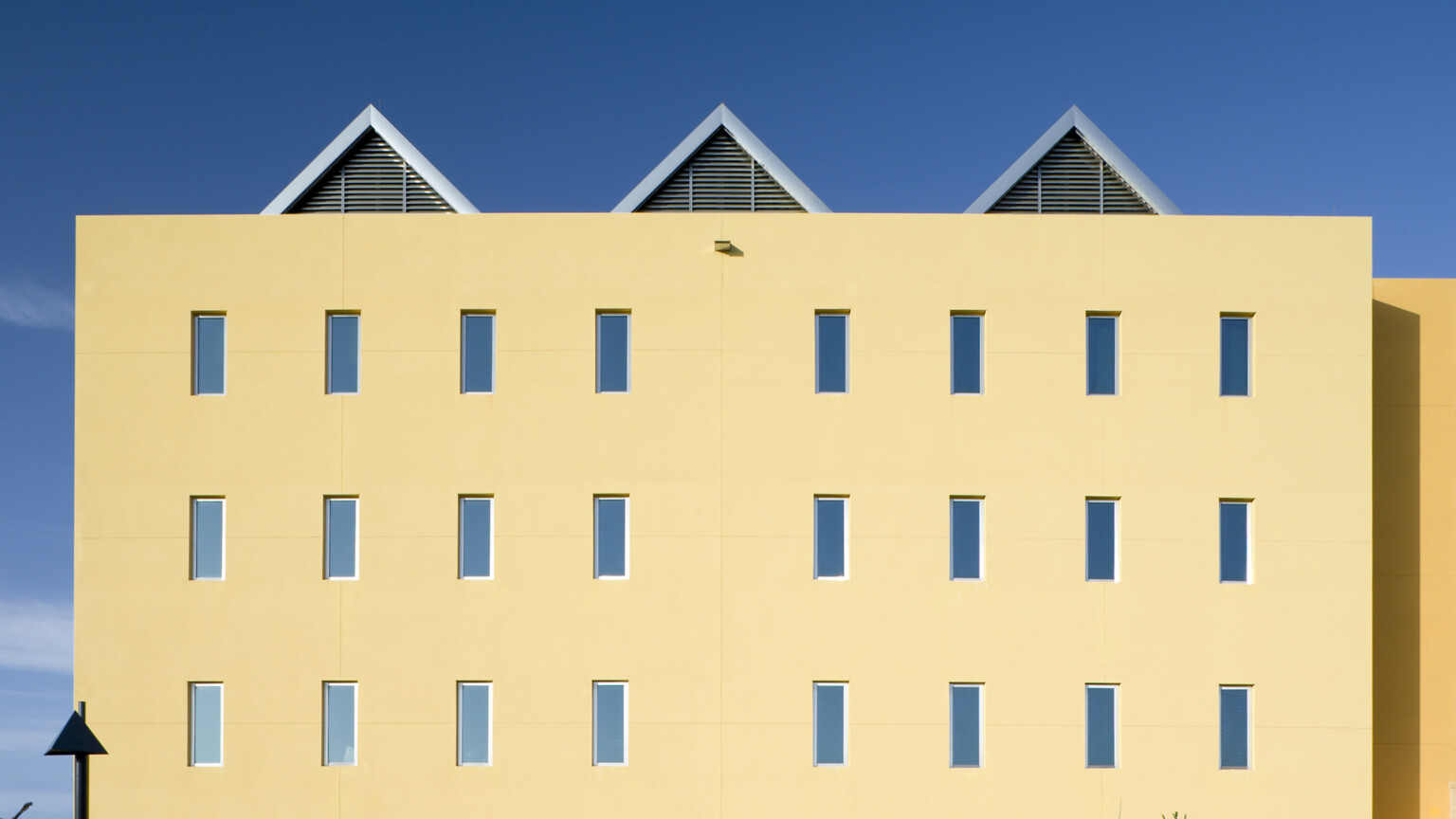 Square yellow building with evenly repeating rectangular windows and palm trees, three peaked roofs spaced evenly on top