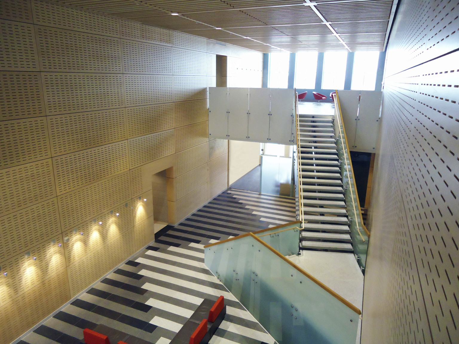 Angled floating stair case with glass side panels extends over wood acoustic panel walls and a black and white tile floor.