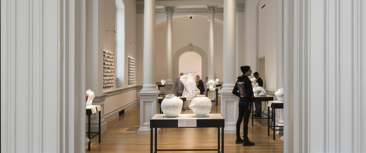 white art gallery with tall pillars displaying pottery on dark tables, framed by crown molding around doorway