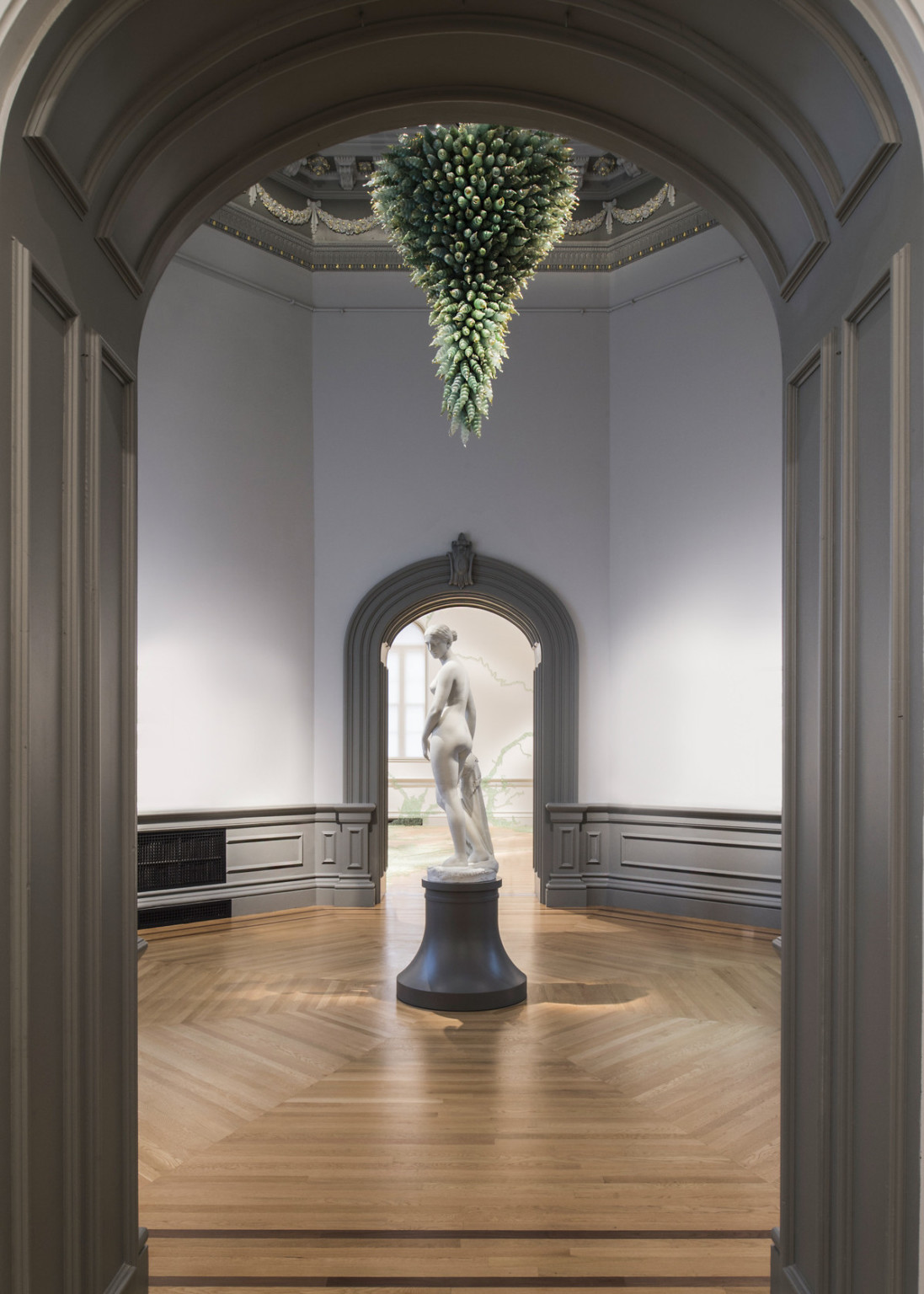 Marble sculpture of woman on black pedestal centered in small room under abstract green sculpture framed by molded door frame