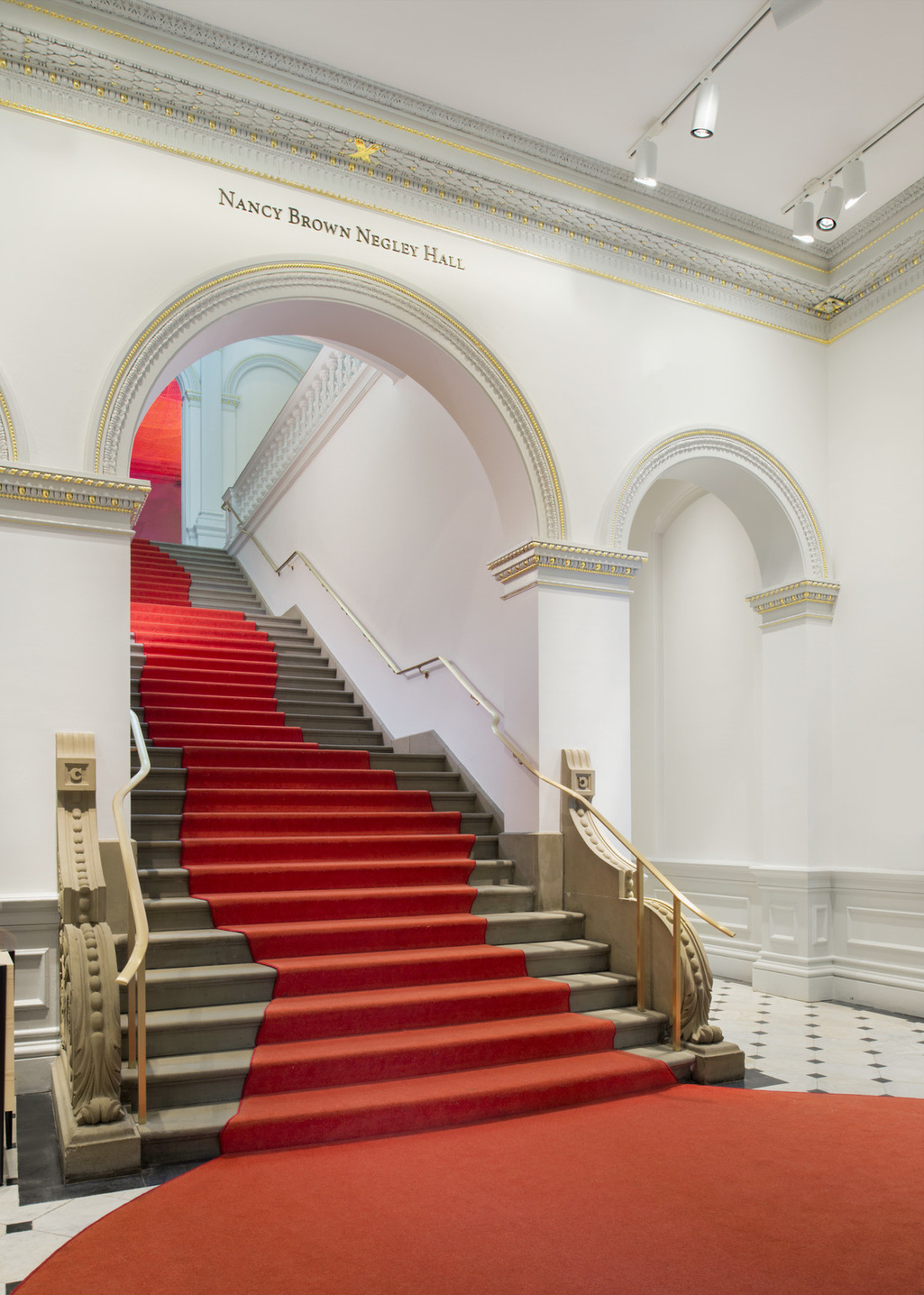 Grand staircase with curved red carpet that widens at base of stairs. Stairs pass through a white arch with gold molding