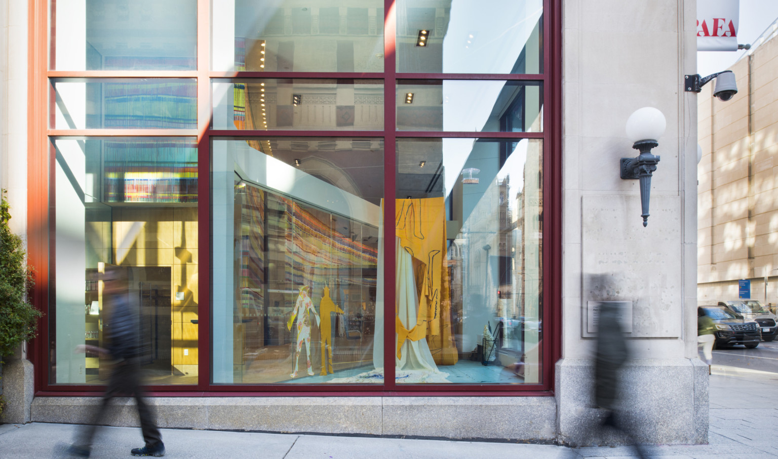 Double height windows with red trim seen from the street next to a light with metal sconce. An art gallery can be seen inside