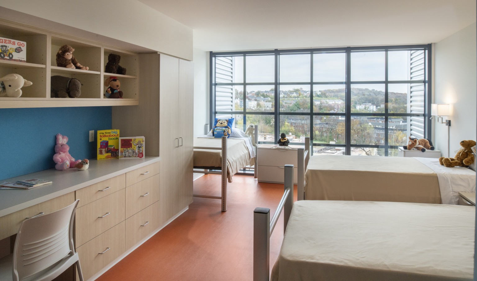 Interior of children's bedroom facing geometric window wall. 3 beds, closets and counter space left with books and toys.