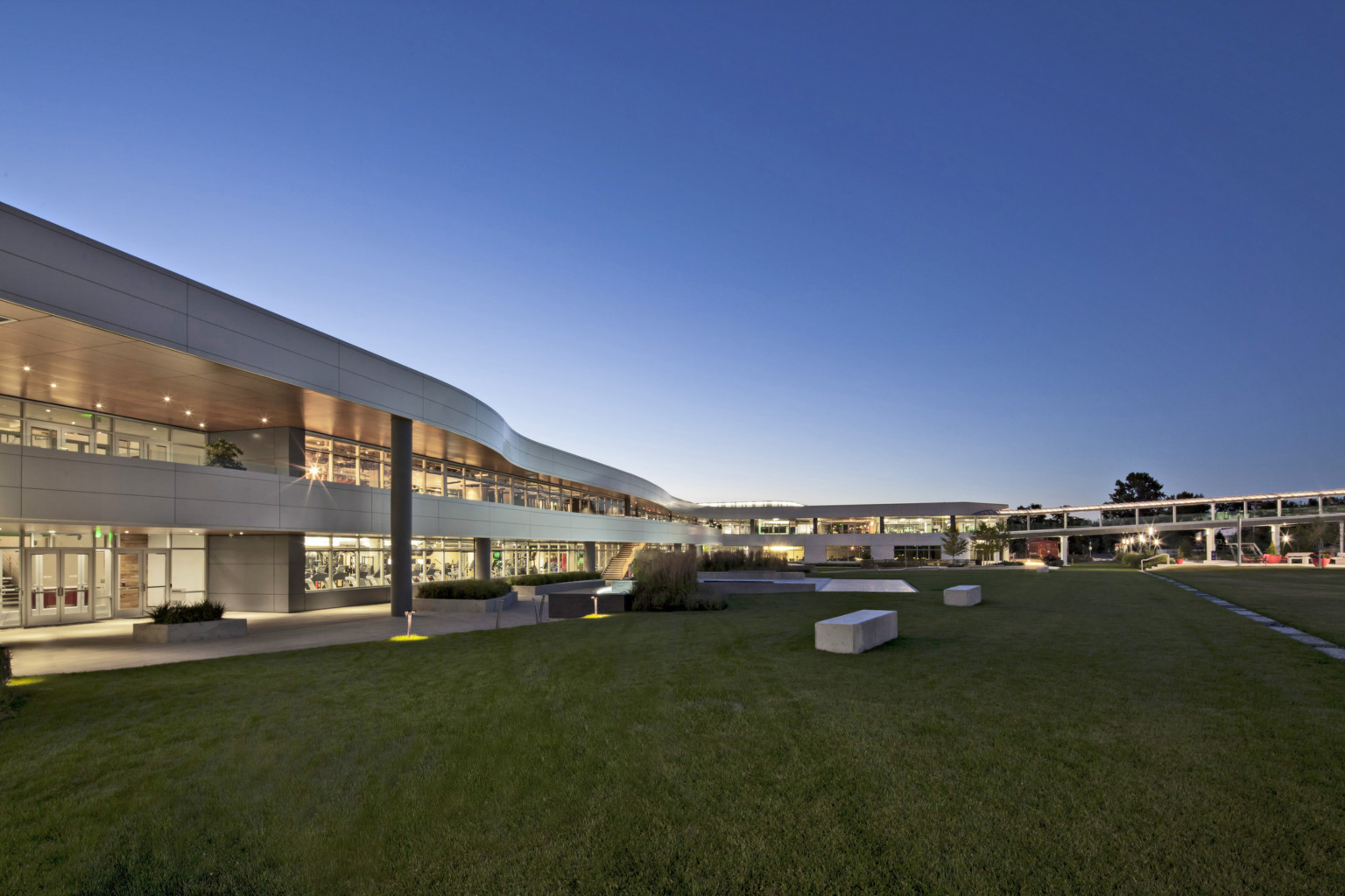 curvaceous silver 2 story campus building with patio and balcony and floor to ceiling windows illuminated at dusk