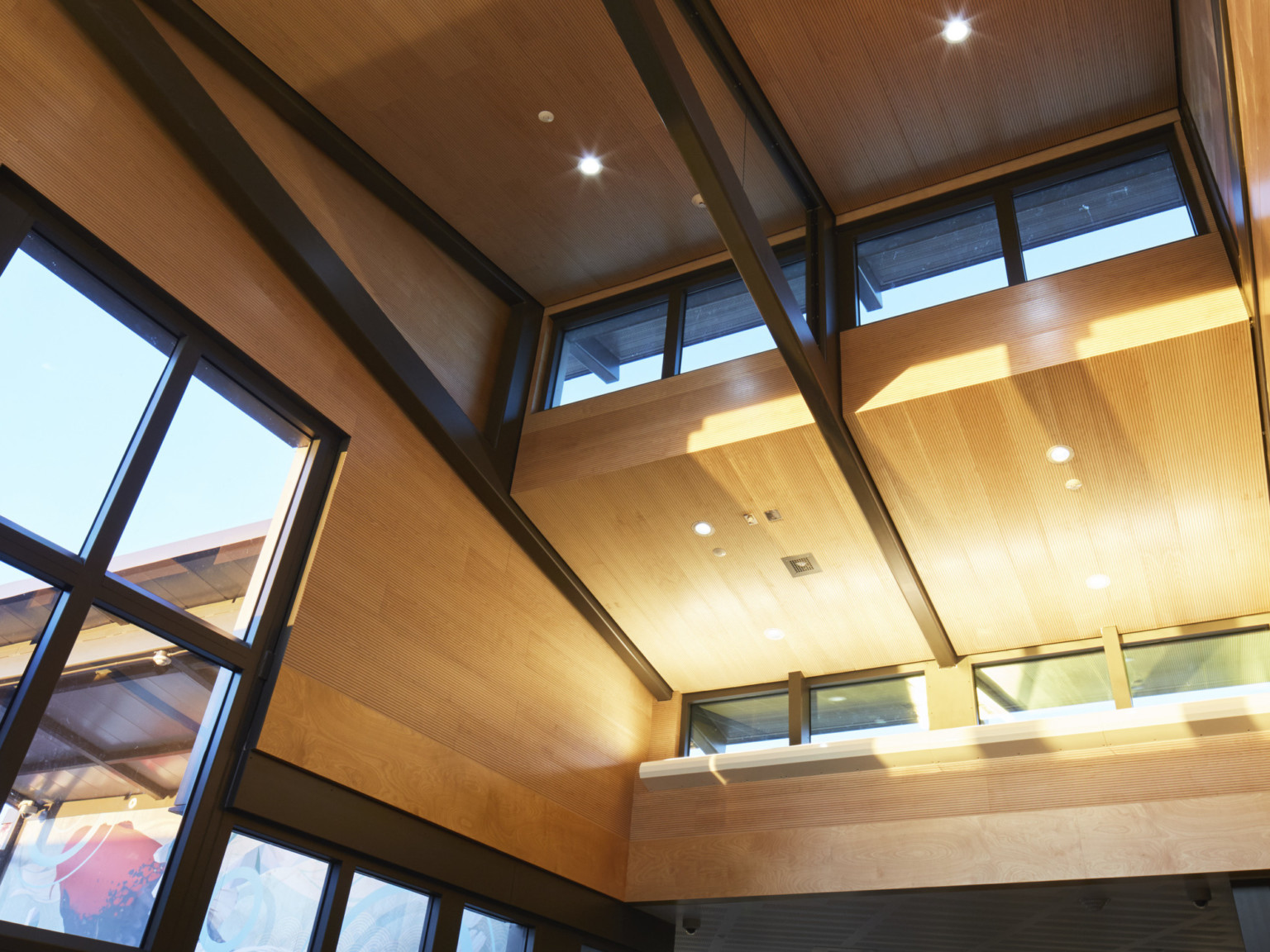 Interior view of double height wood panel hallway with large black framed windows and black trusses on drop ceiling detail