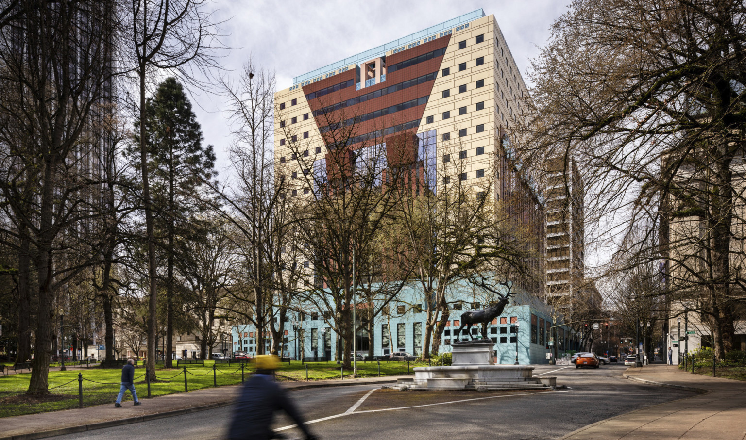 View of beige building with red angular stripe, blue ribbon design at side, over a teal multistory base, surrounded by trees