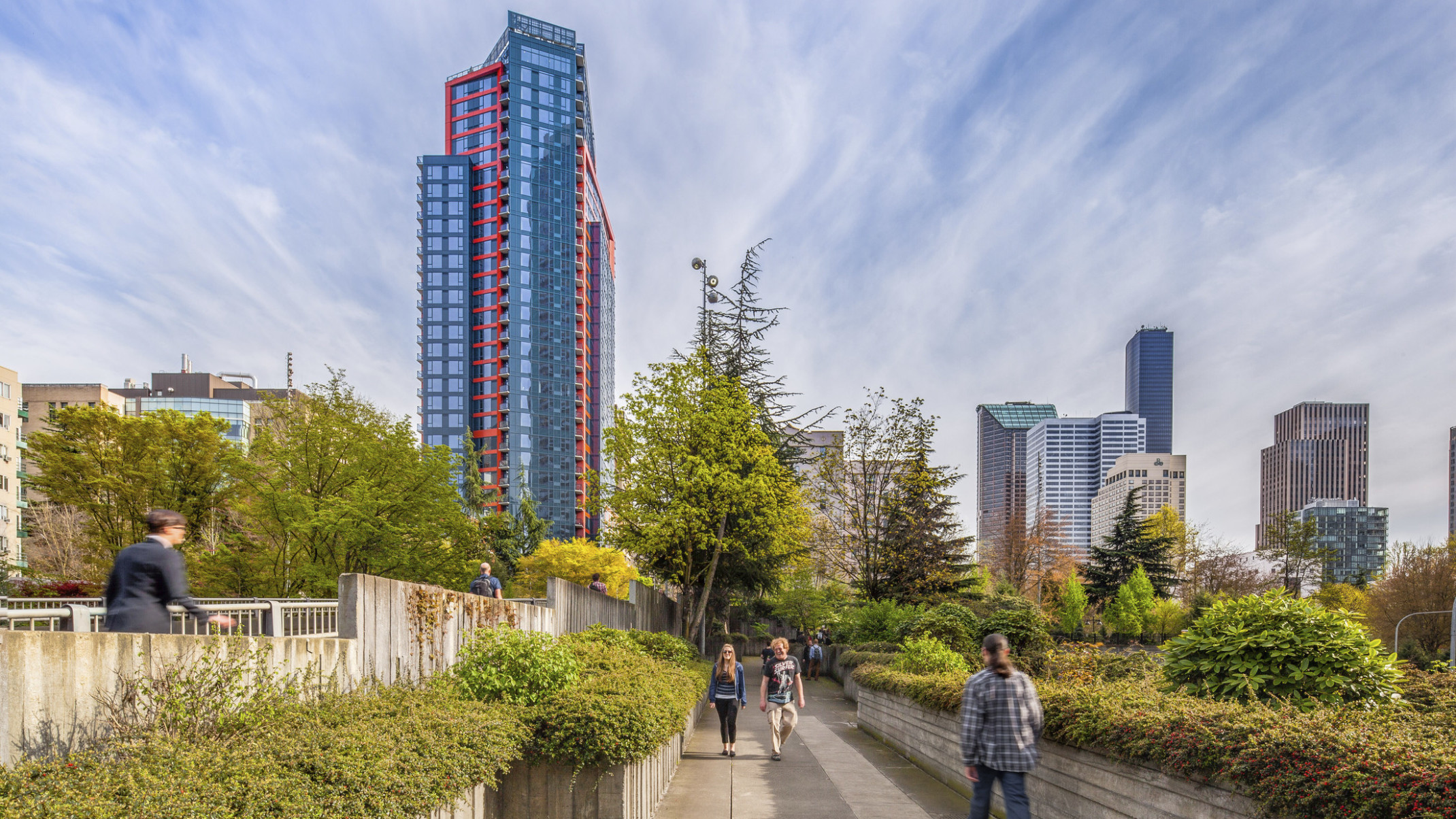 Cielo, a layered glass building with red and blue wrapping facade, behind a tree lined walkway surrounded by skyscrapers