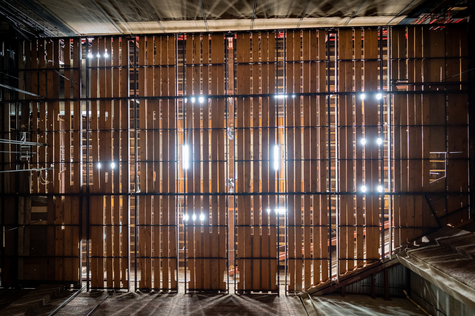 Wood panel wall sections with large spaces between planks and sections, light shining through in room with raw floor