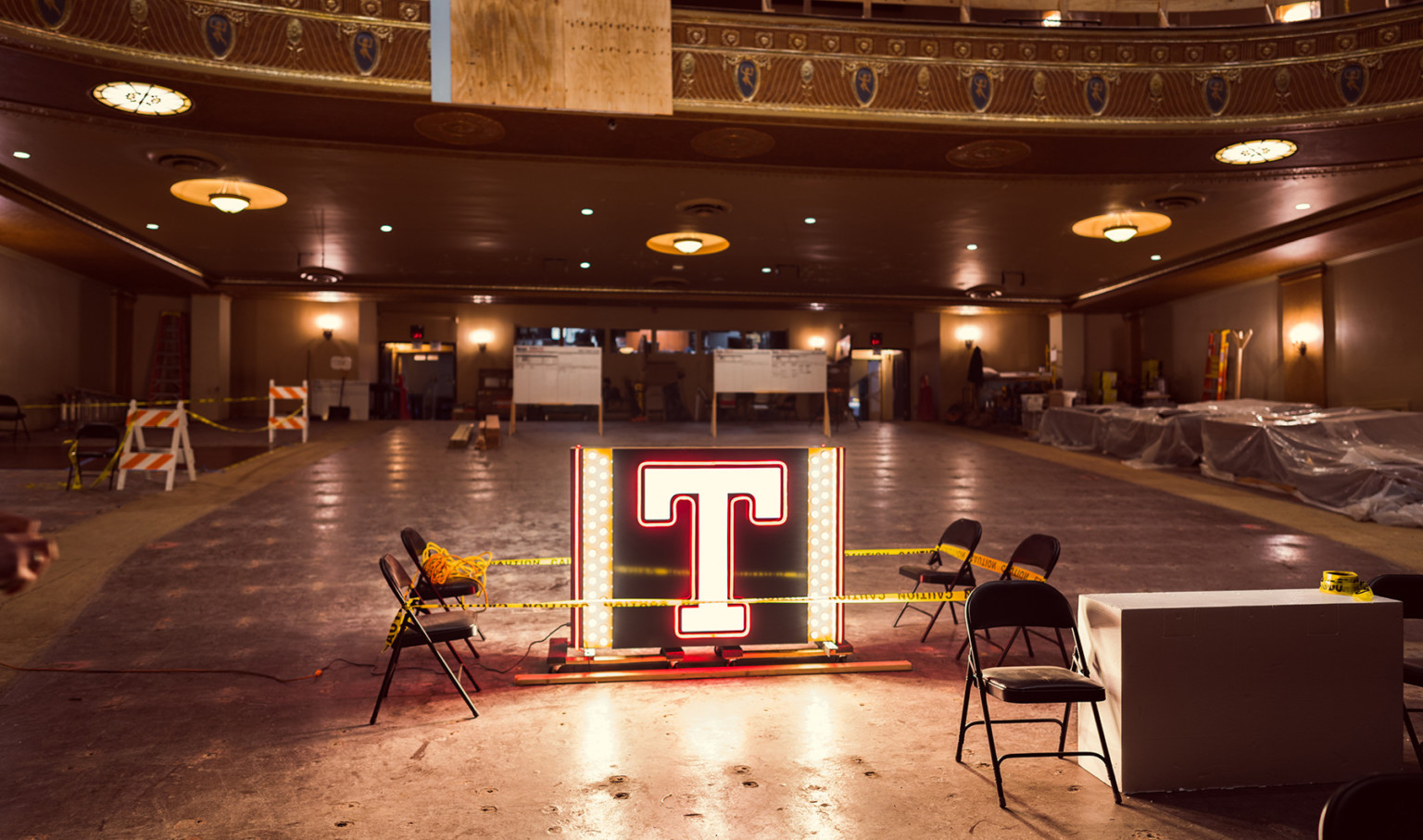 Illuminated letter T in box with lights on sides inside of partially constructed theater with no seats, molding on balcony