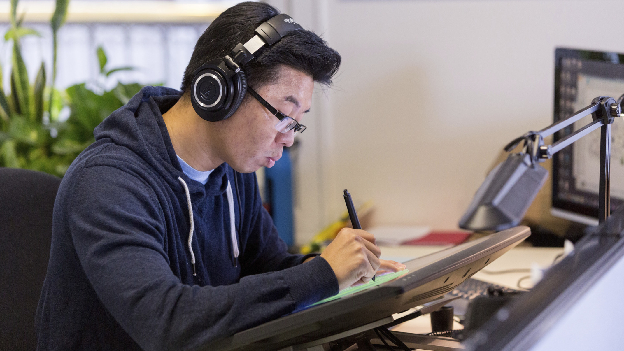Man sketching on touchscreen with headphones on at desk in white office. Plants in window behind