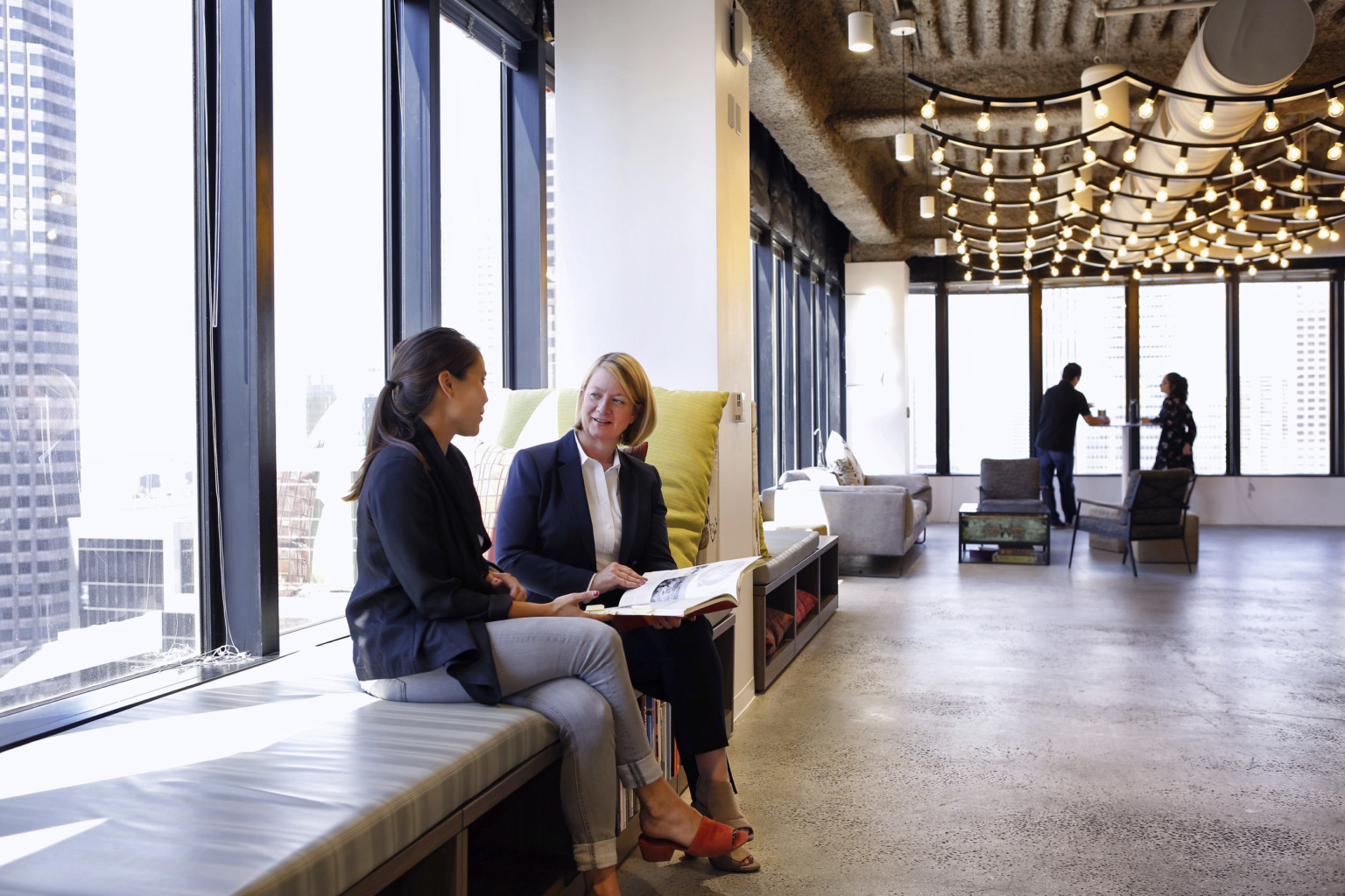 Cushioned window seat against wall of floor to ceiling glass. Open ceiling has bulb lights strung vertically over ductwork.