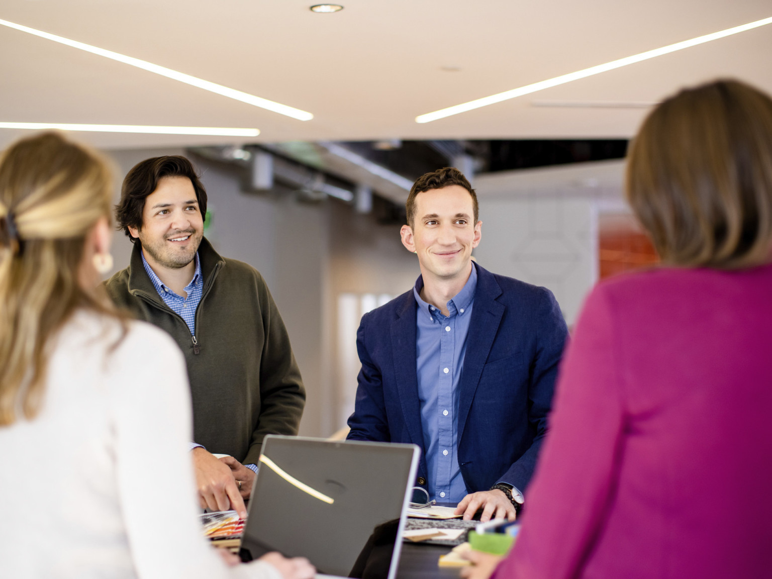 a group of smiling young professionals collaborating around a standing height table under a ceiling with diagonal lighting