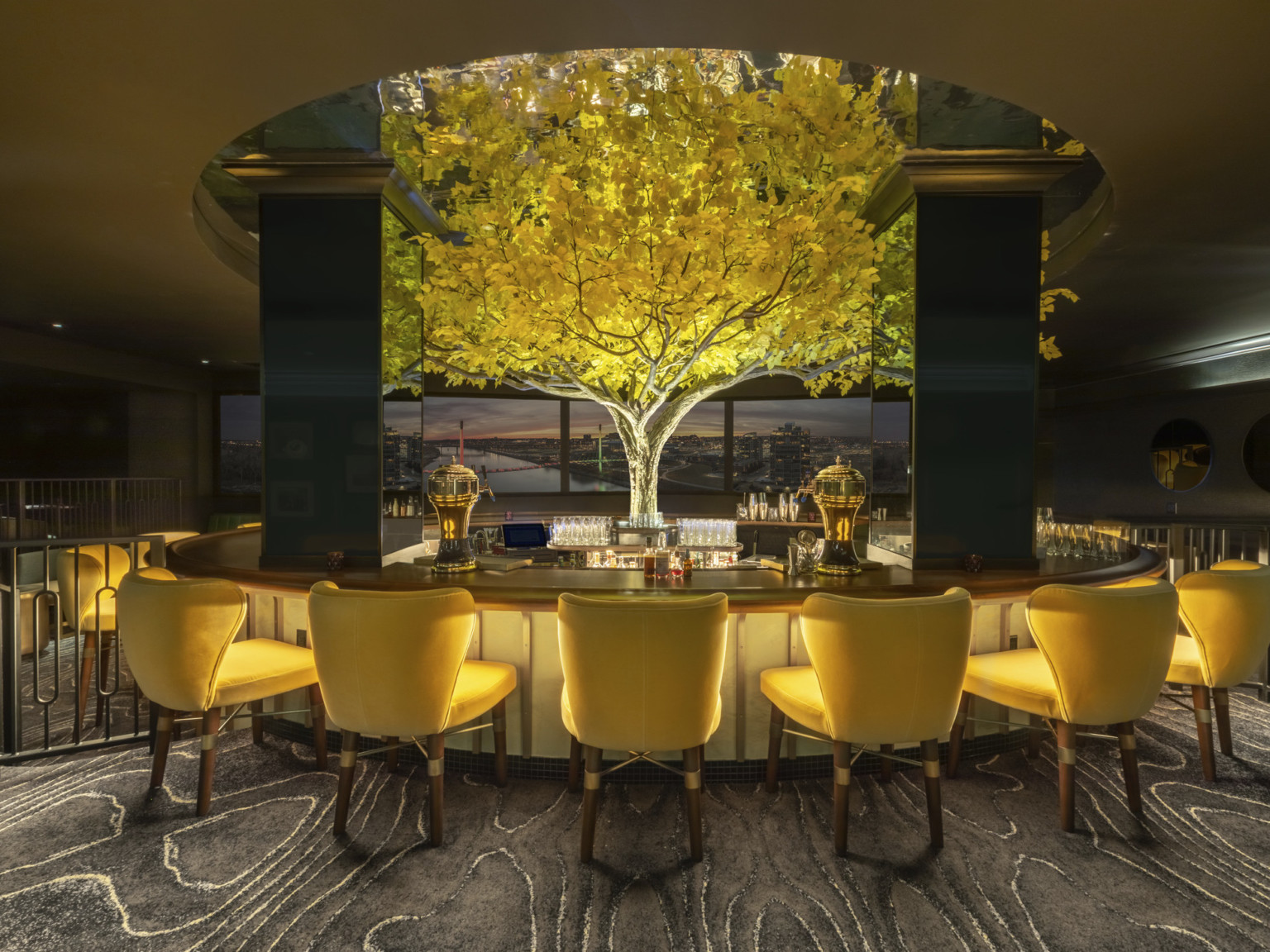 an illuminated yellow tree in the midst of in the round high top bar with yellow seats. Tree rises above round ceiling cutout