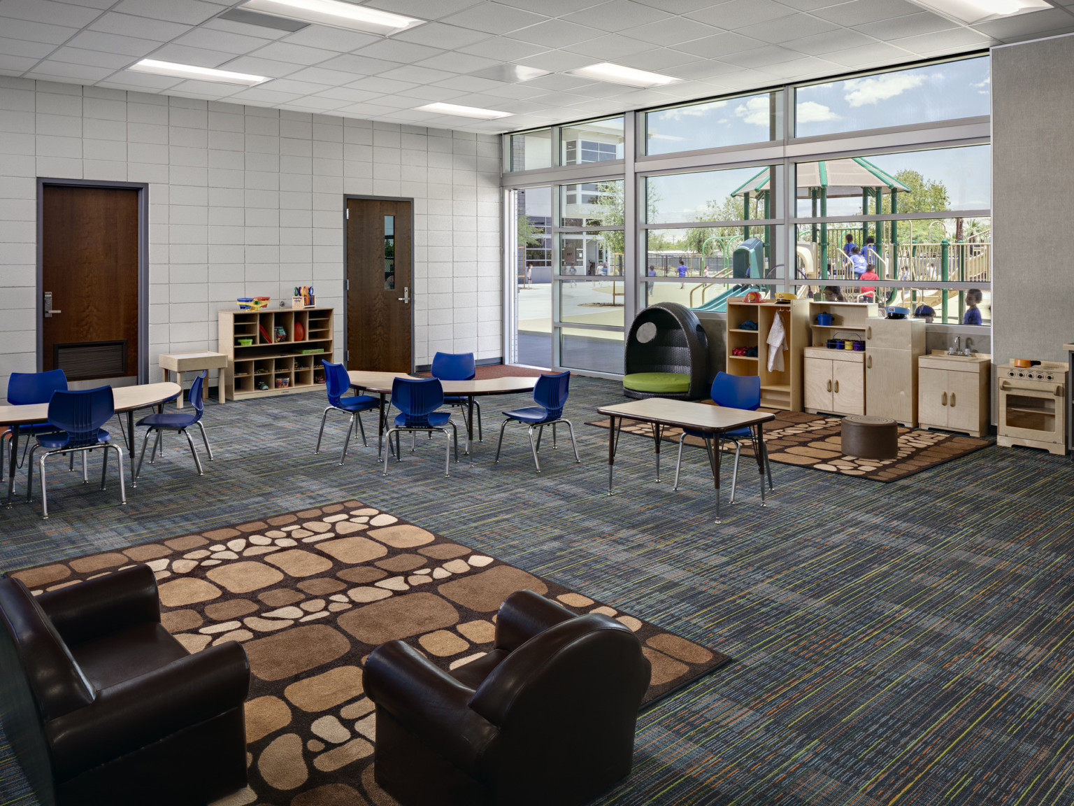 White classroom with children's tables and chairs, brown patterned rug with comfortable chairs, and floor to ceiling windows