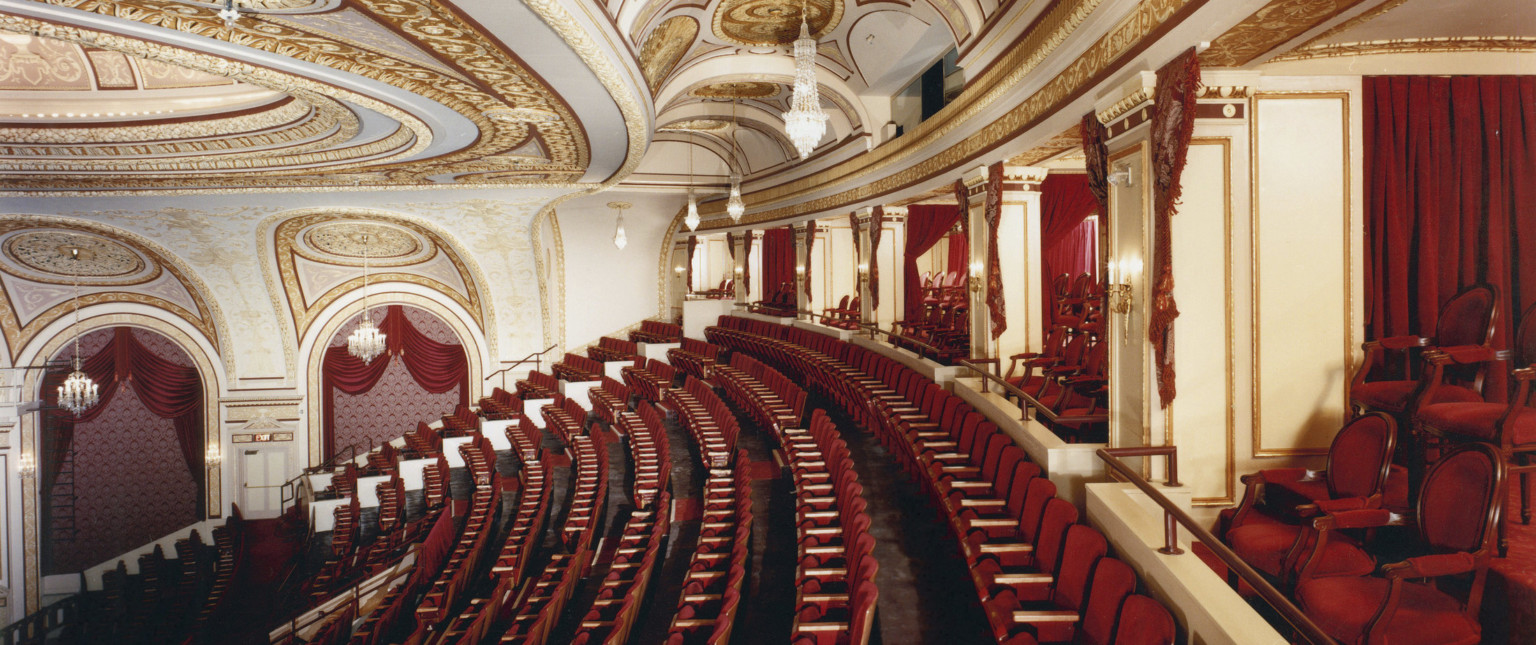 Box seats and back rows of audience in the Connor Palace Theater, with red seats and white walls with gold scroll details