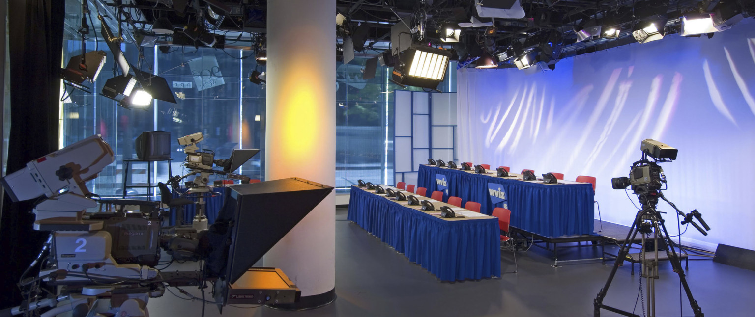 Television studio at street level with three cameras pointed at two rows of tables with chairs and phones, lit from above