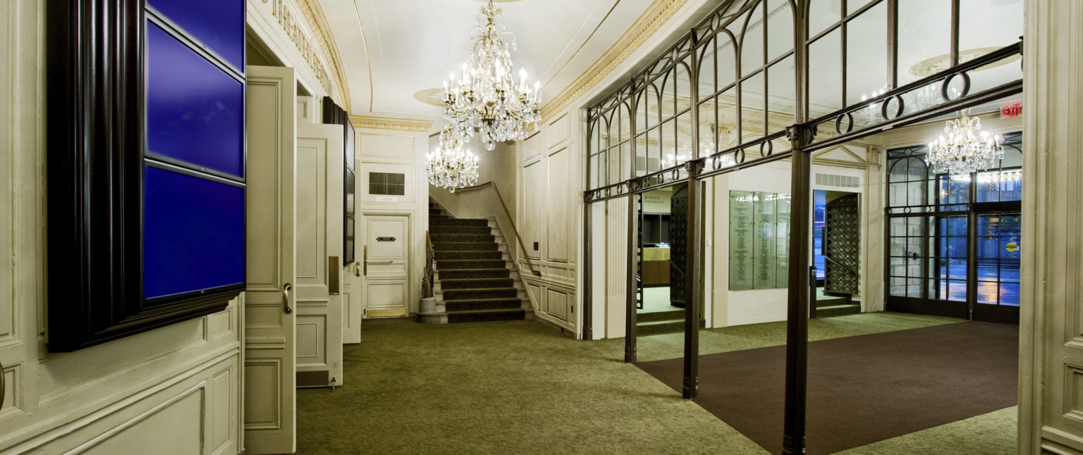 Green carpeted hallway leading to stairs, white walls with doors left and chandeliers hang from above framed by gold molding