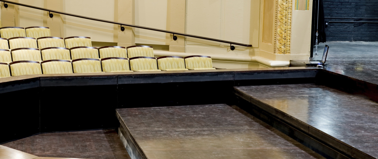 Thrust style proscenium with three black steps leading up to the stage in front of cream colored audience seats
