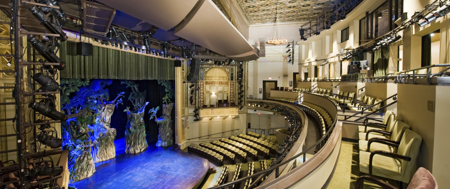 Multiple balconies in a theater with yellow seats and dark details, box seat center with arch overhang and gold details