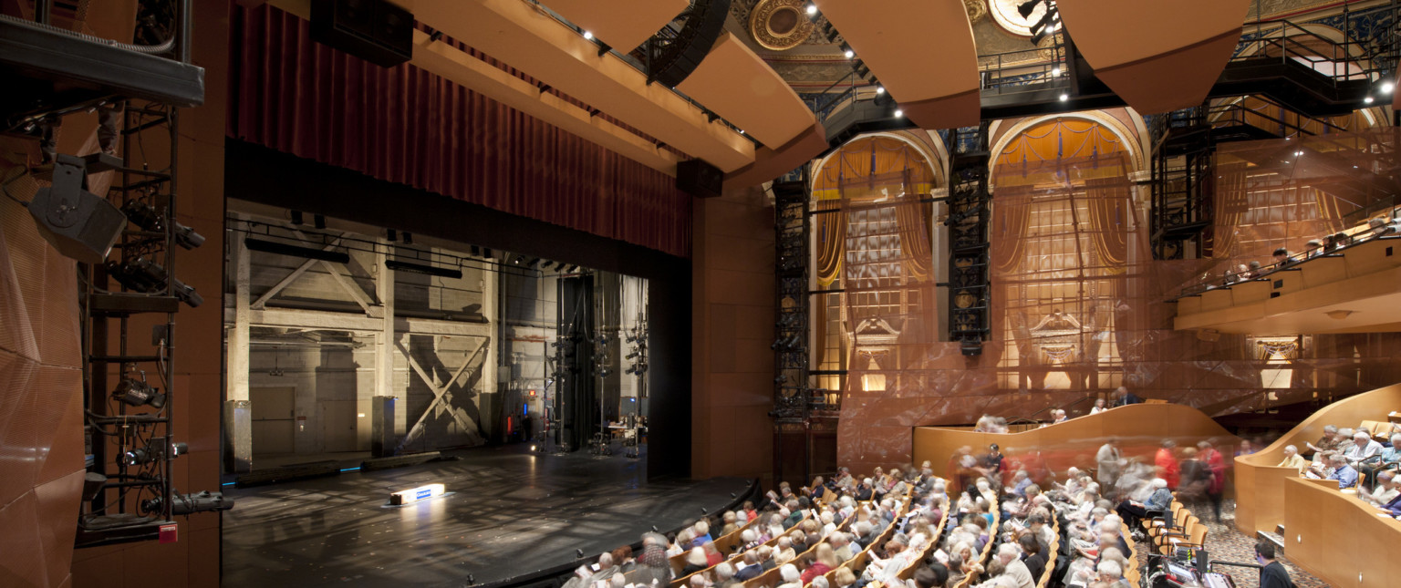 Theater seen from the side over the audience with large arch shaped windows. The stage is empty and stage right can be seen