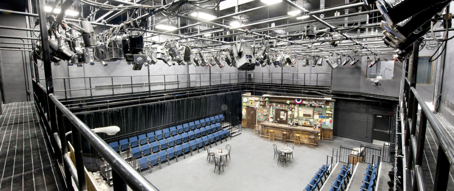 Black box theater with side seating, lights hang on a metal grid above. The stage is set with tables, chairs, and a bar