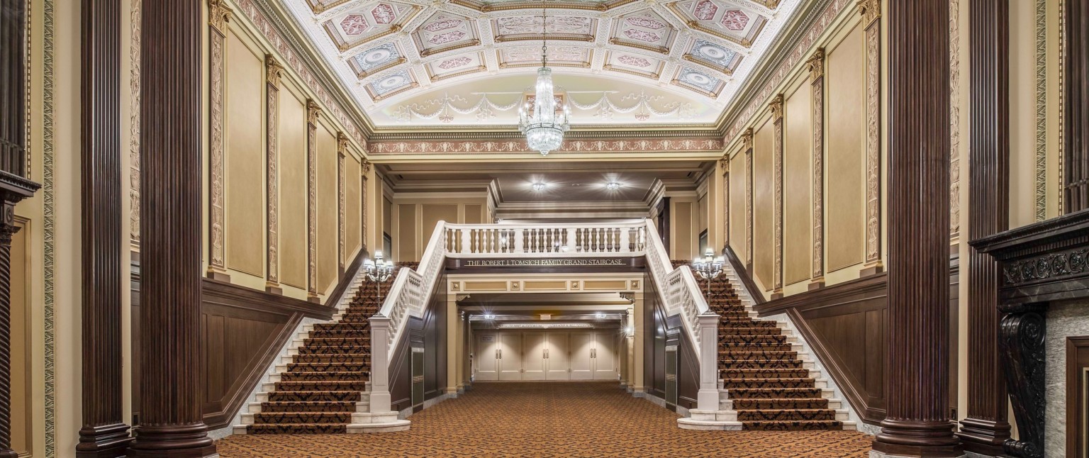 Grand staircase carpeted beneath ornate multicolored coffered ceiling with chandelier framed by ribbed pillars