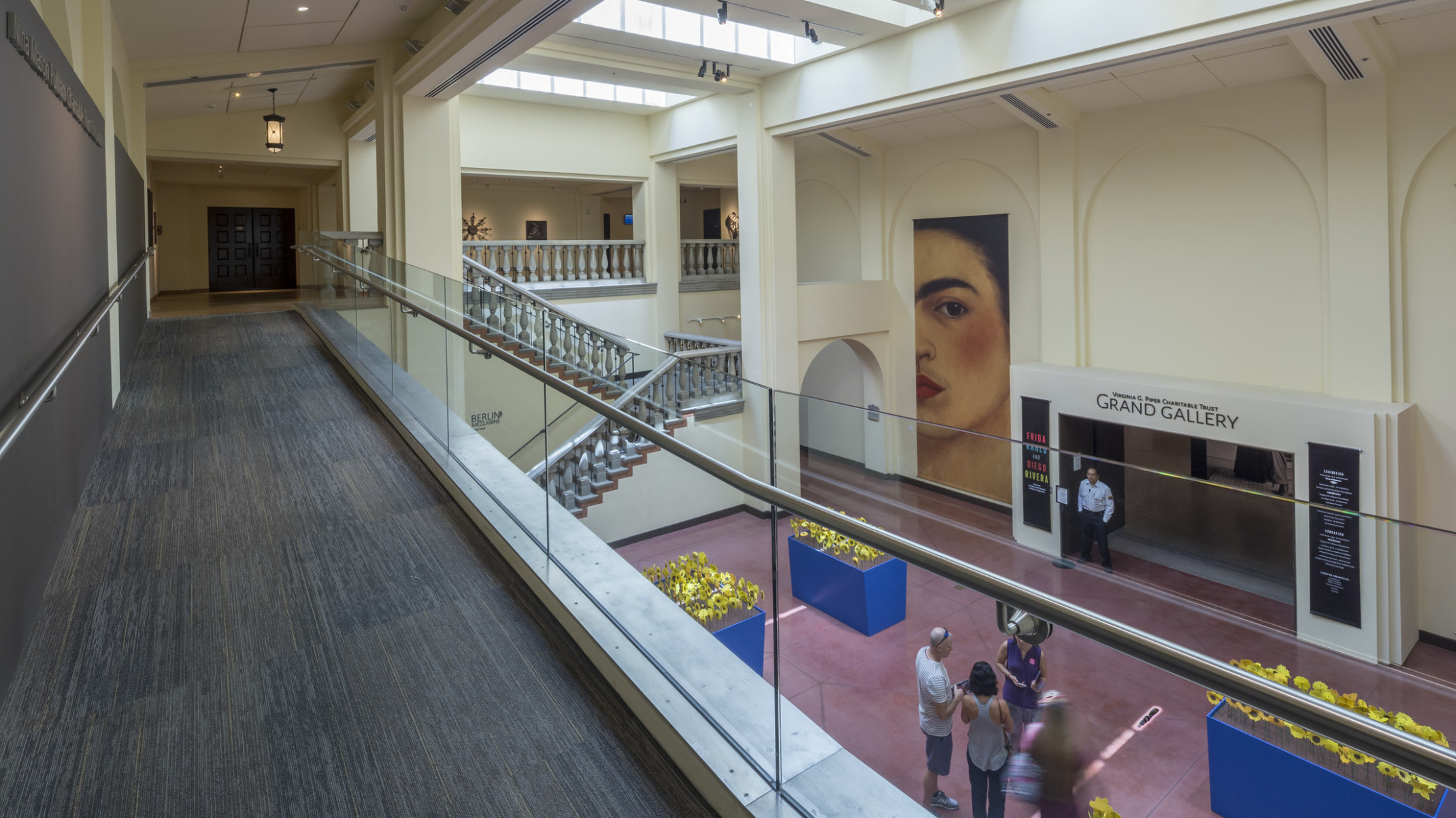 A carpeted walkway overlooks a staircase and an entryway labeled Grand Gallery in cream colored room with skylights