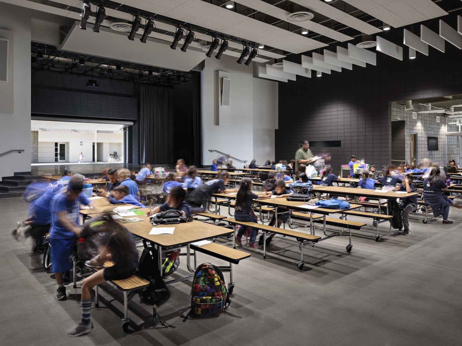 Double height room with long wood lunch tables in front of stage with back wall opened to hallway. Grey acoustic panels above