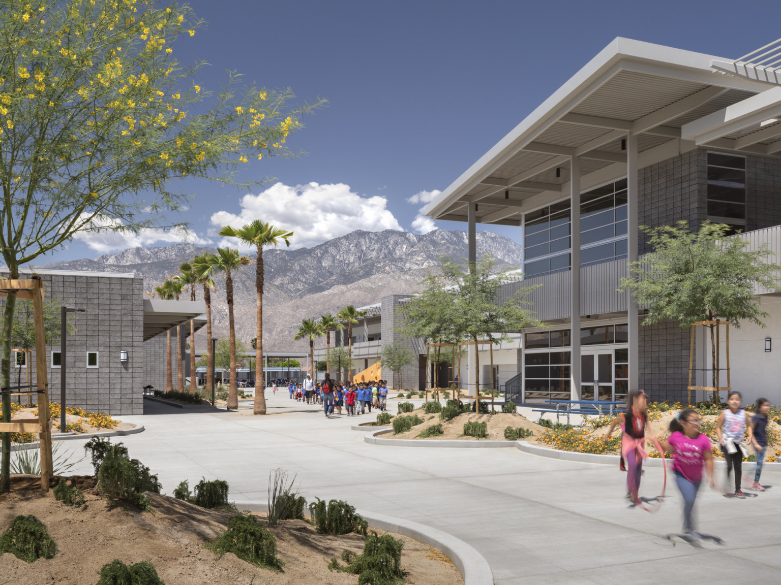 courtyard with palm trees between grey stone buildings with silver canopy, Santa Rosa and San Jacinto Mountains seen beyond