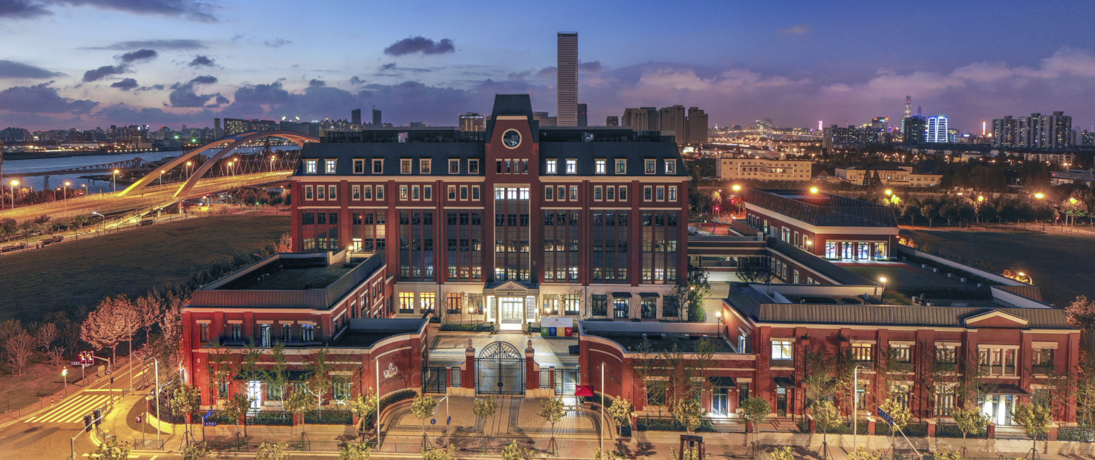 View of brick school with dark stone roof. A central tower extends above entry of the main building, illuminated at night.