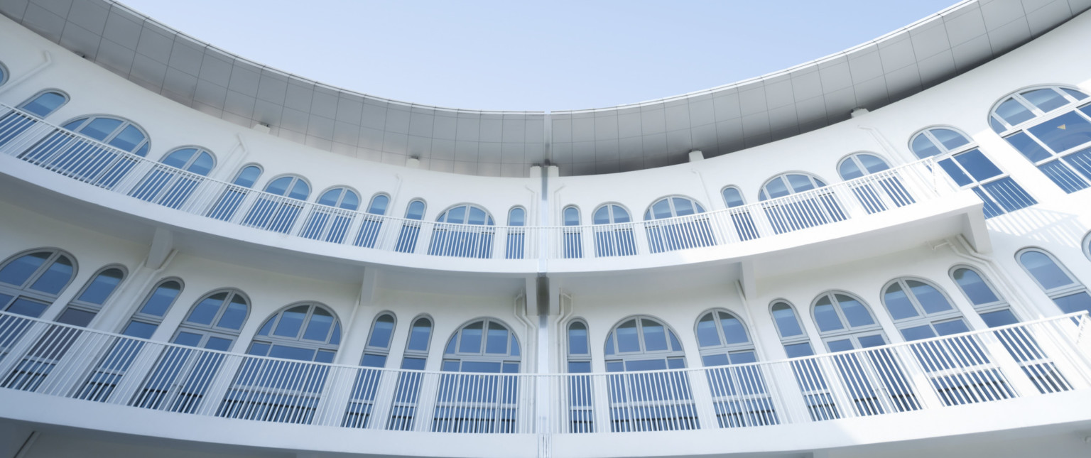 View looking upwards at curved white facade with railed exterior walkways along lines of arched windows. Tiled white canopy