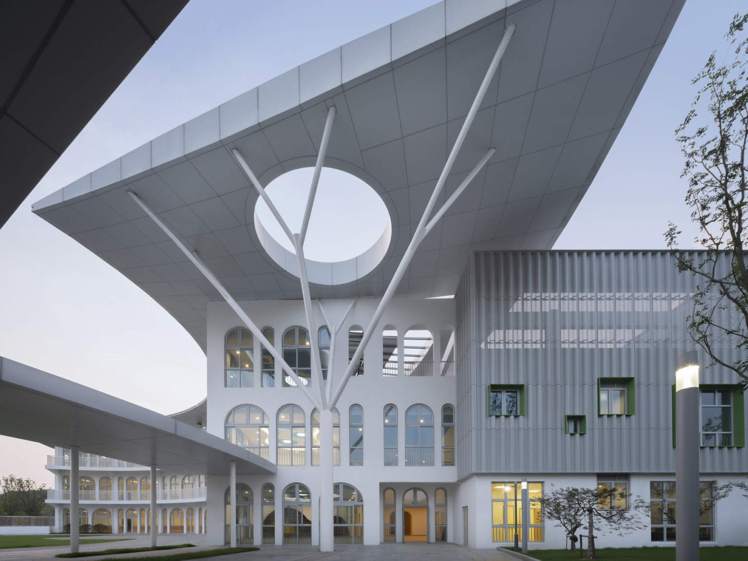 Walkway to white building with arched windows under angular canopy with circular cutout supported by branching white column