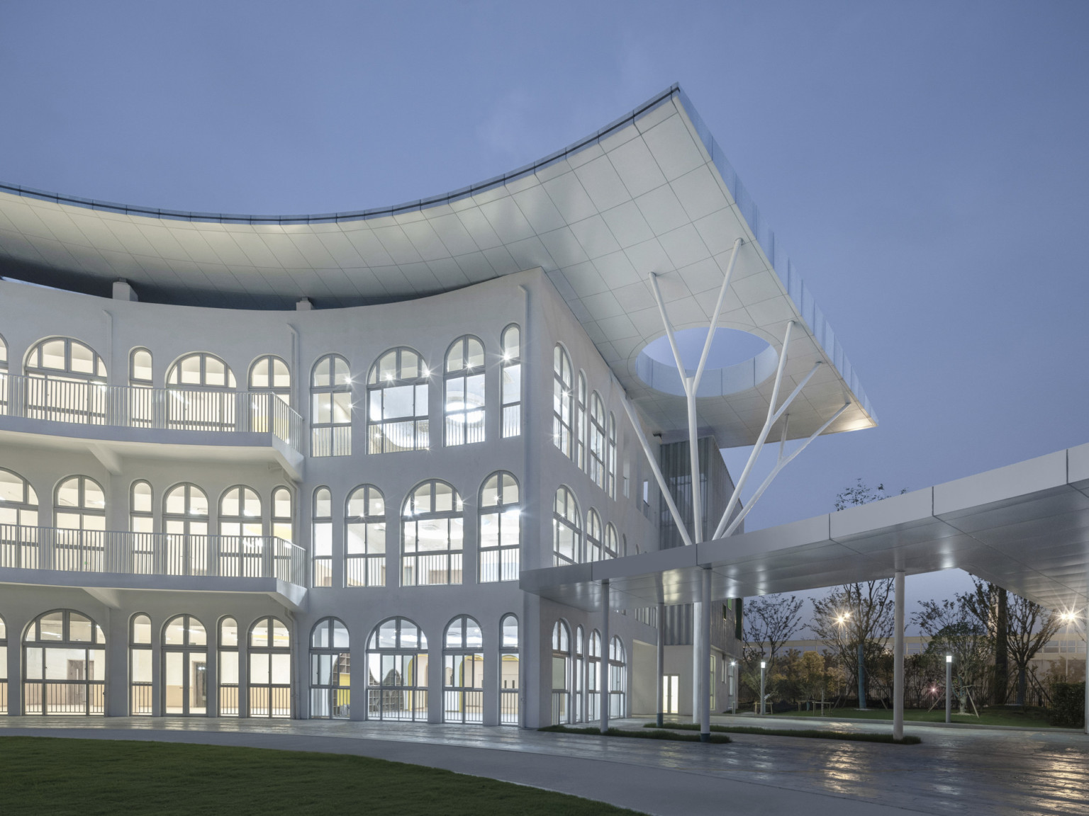 White 3 story building illuminated in evening with tiled overhang to right with circular cutout supported by branching beams