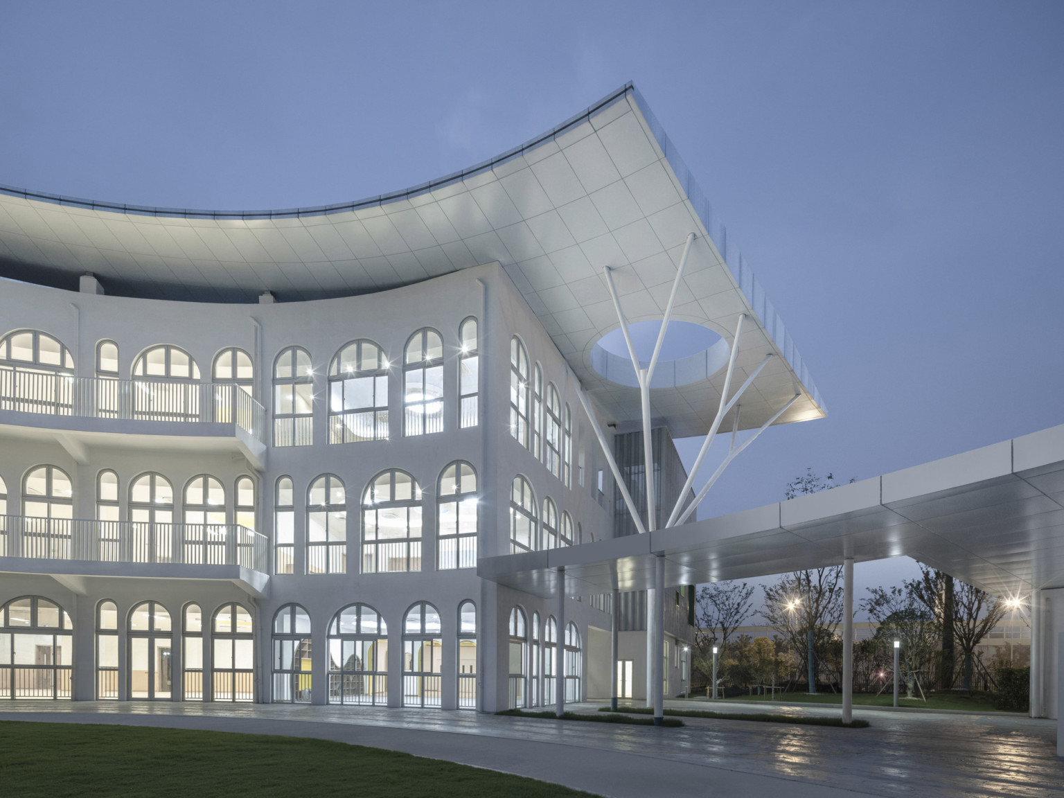 White 3 story building illuminated in evening with tiled overhang to right with circular cutout supported by branching beams