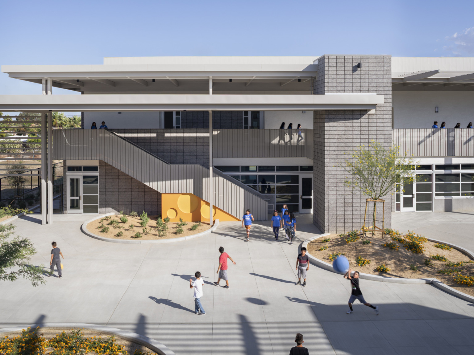 Courtyard in front of 2 story stone building. Orange accent wall with round cut outs. Stairs in front to 2nd floor walkway