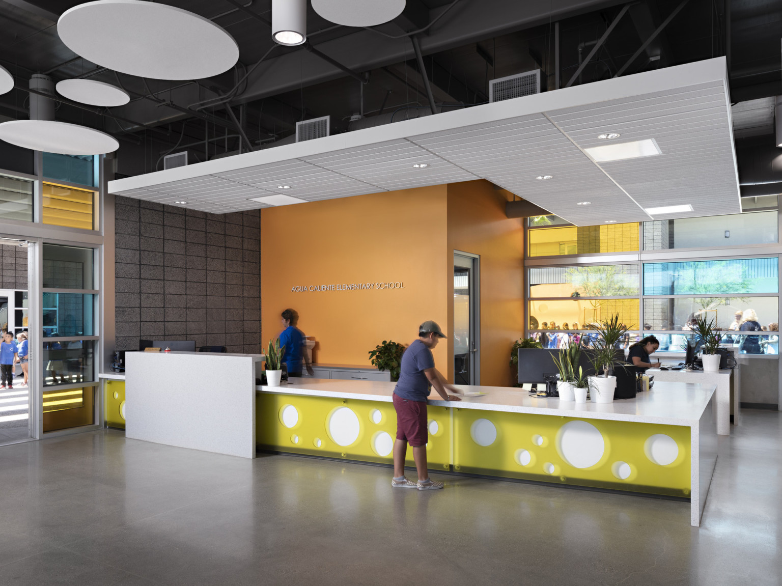 Reception desk with Agua Caliente Elementary School sign. White desk has yellow overlay with round cutouts, colored windows