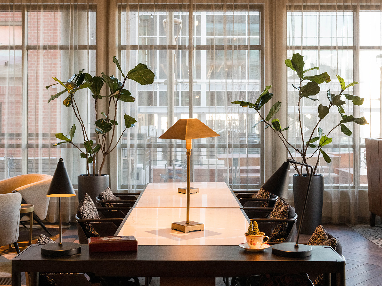 gold and black table lamps on a glossy white table lined with chairs. Floor to ceiling windows have closed sheer curtains.