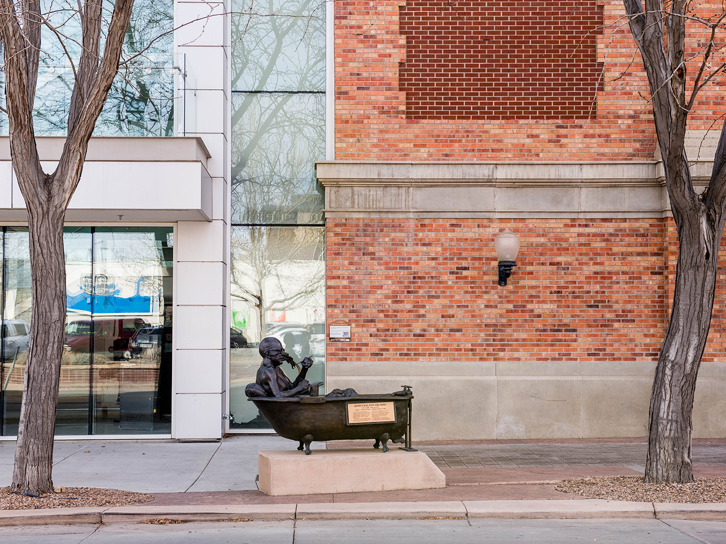 bronze sculpture of a man in a bathtub on a plinth on the sidewalk's edge between two trees in front of glass, steel, brick