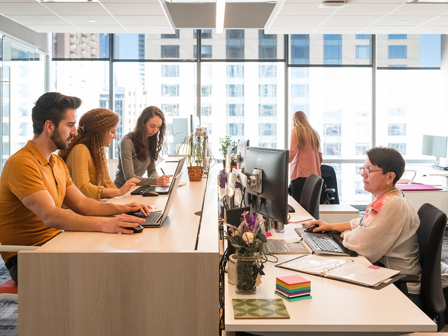 standing height work desk adjacent to sit stand open benching style desks in front of floor to ceiling windows with 5 people