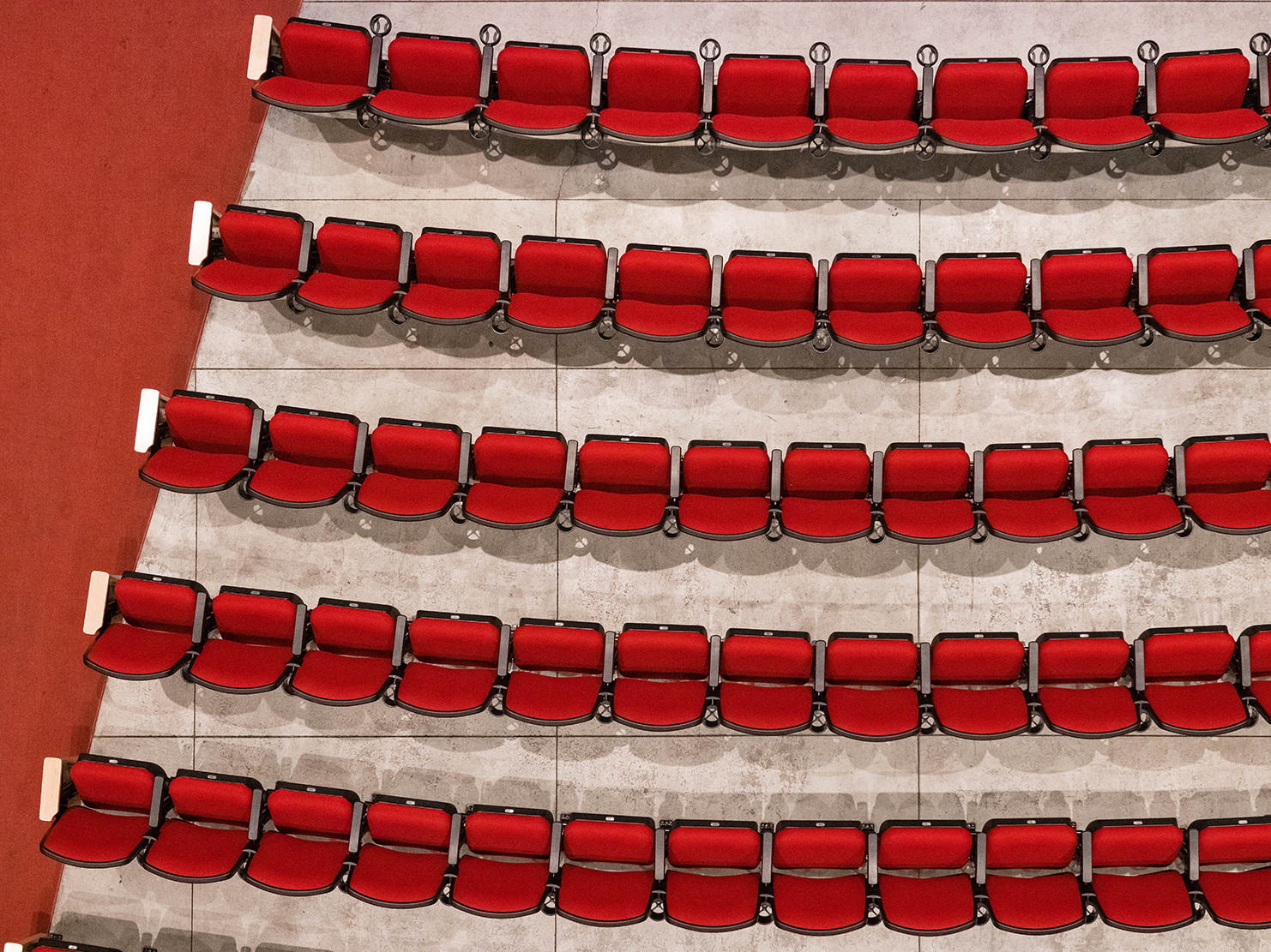 five continuous curved rows of red soft upholstered theater seats on gray concrete floors. a red carpet runs next to seats