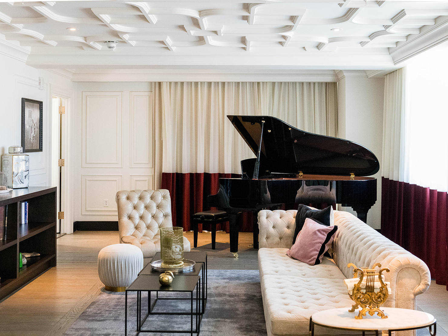 white molding on ceiling and walls, sheer white and crimson velvet curtains, with a glossy piano behind cream tufted seating