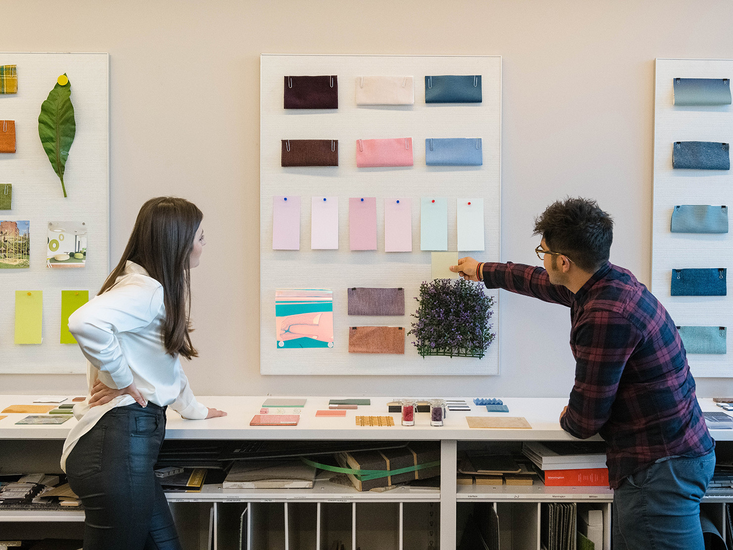 palette boards on walls with various color samples, inspiration images, and flowers and leaves pinned up. two people look on